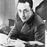 Albert_Camus,_gagnant_de_prix_Nobel,_portrait_en_buste,_posé_au_bureau,_faisant_face_à_gauche,_cigarette_de_tabagisme